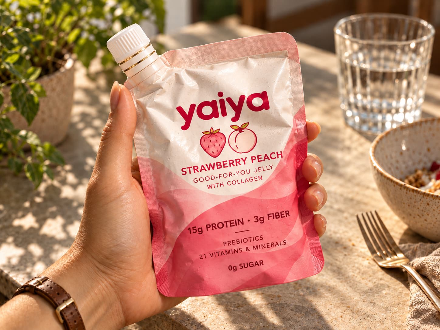 A hand holding a Strawberry Peach pouch at a sunny table, beside a nearly finished bowl, a fork and a glass of water.